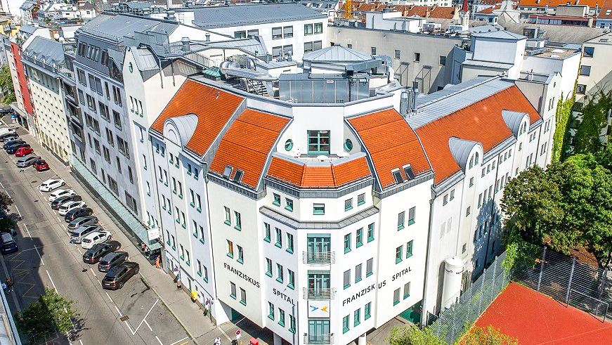 Luftbild des Franziskus Spitals Margareten in Wien: weißes Eckgebäude mit rotem Ziegeldach, Aufschrift „Franziskus Spital“, angrenzende Straße mit parkenden Autos und Sportplatz mit rotem Boden.